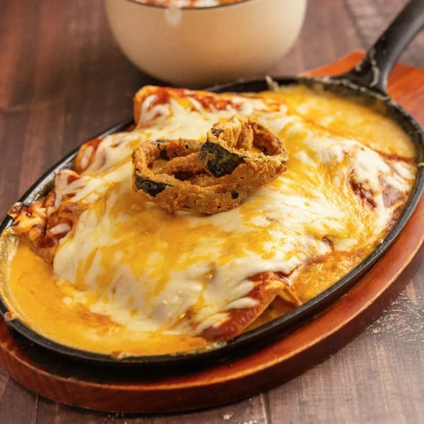 Sizzling skillet of cheesy enchiladas topped with crispy fried onion rings on a wooden table.