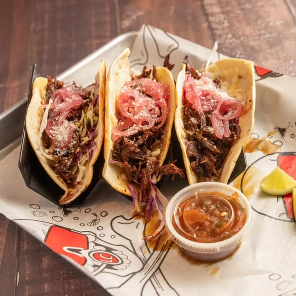 Three tacos with shredded meat, pickled red onions, and cheese, served with salsa and lime wedges.