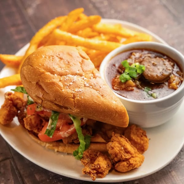 A fried shrimp po'boy with fries and a bowl of gumbo on a white plate.