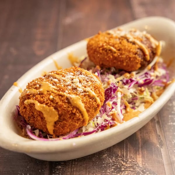 Two crispy, breaded croquettes drizzled with sauce and sprinkled with cheese, served on a bed of coleslaw.
